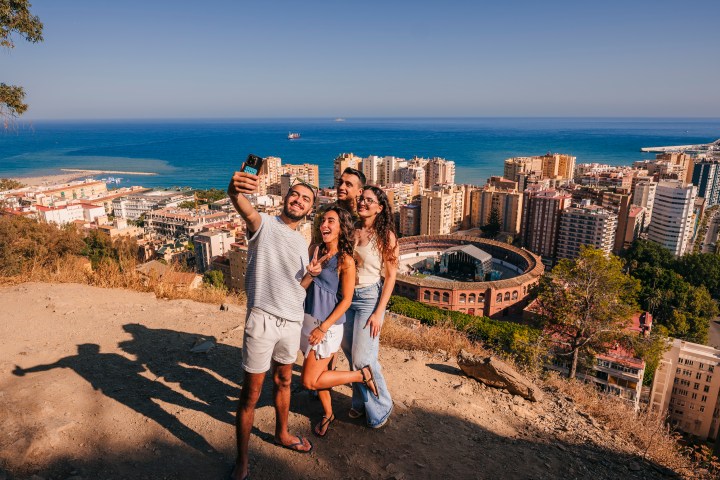 Four people taking a selfie on a hill with a cityscape and ocean in the background.