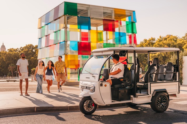 Group of people walking near a colorful glass cube structure and an electric tuk-tuk in daylight.