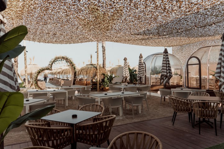 Outdoor beach cafe with tables, chairs, and striped umbrellas under a camouflage canopy.