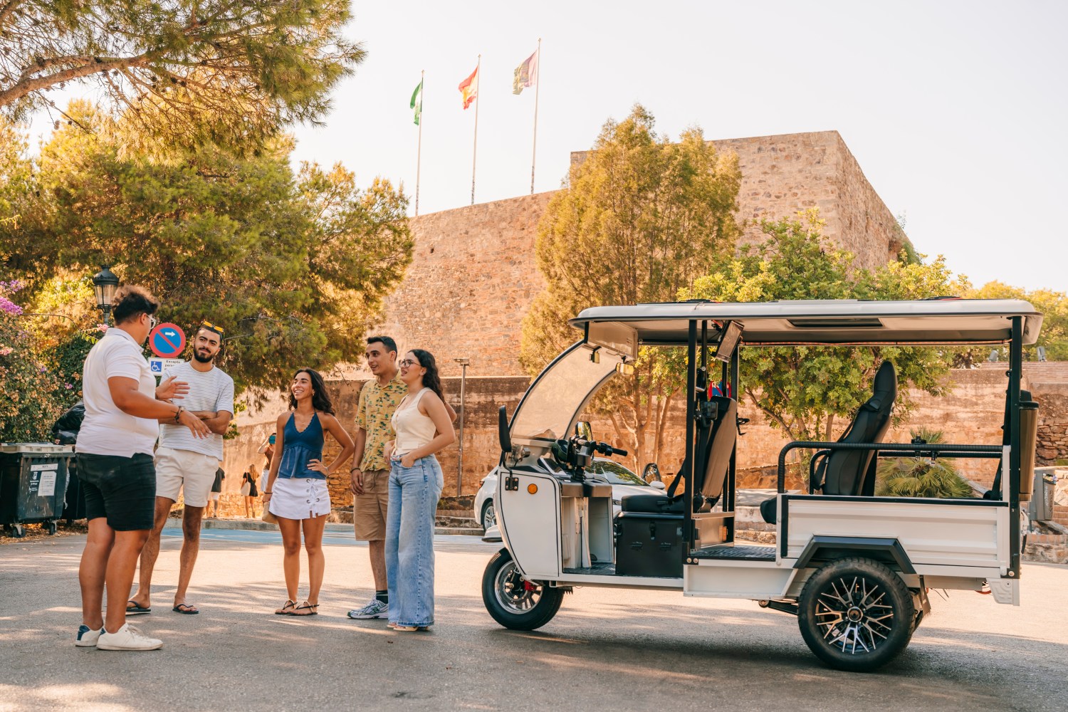 Guía turístico explicando el tour a un grupo de jóvenes junto a un tuk-tuk eléctrico frente a la muralla de la Alcazaba de Málaga