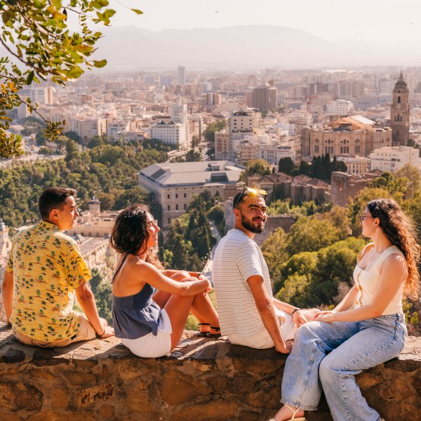 Four people sitting on a stone wall overlooking a cityscape with historic buildings and trees.