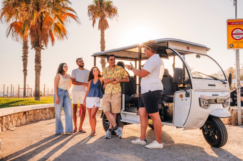 Group of people standing by a parked tuk-tuk near palm trees at sunset.