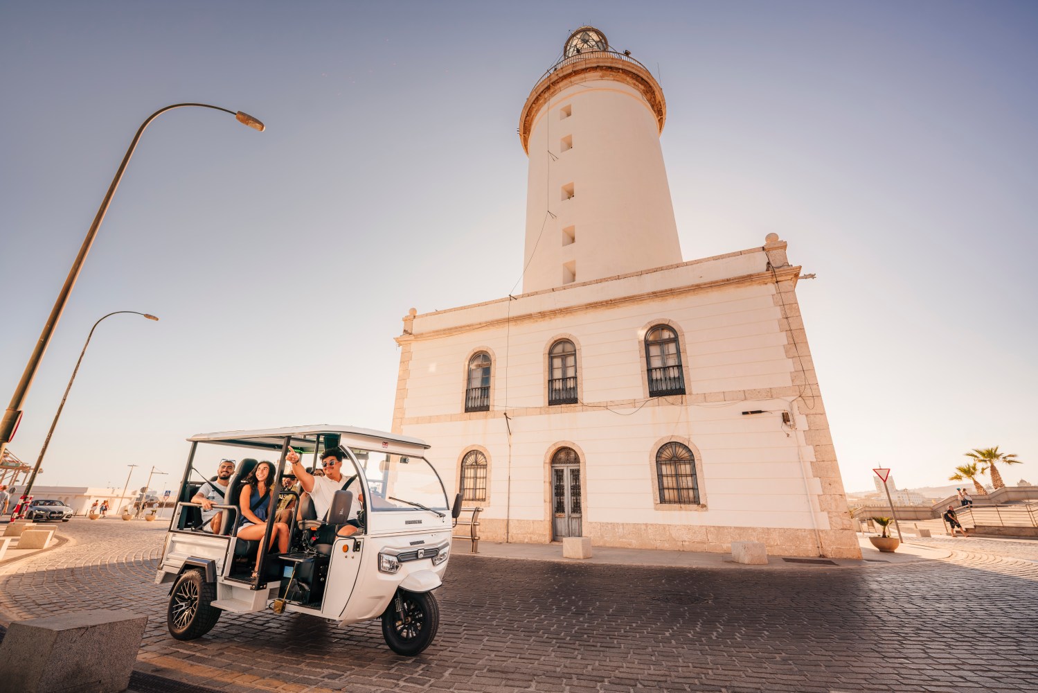 Tuk-tuk turístico con pasajeros frente a La Farola de Málaga en el puerto durante un día soleado.