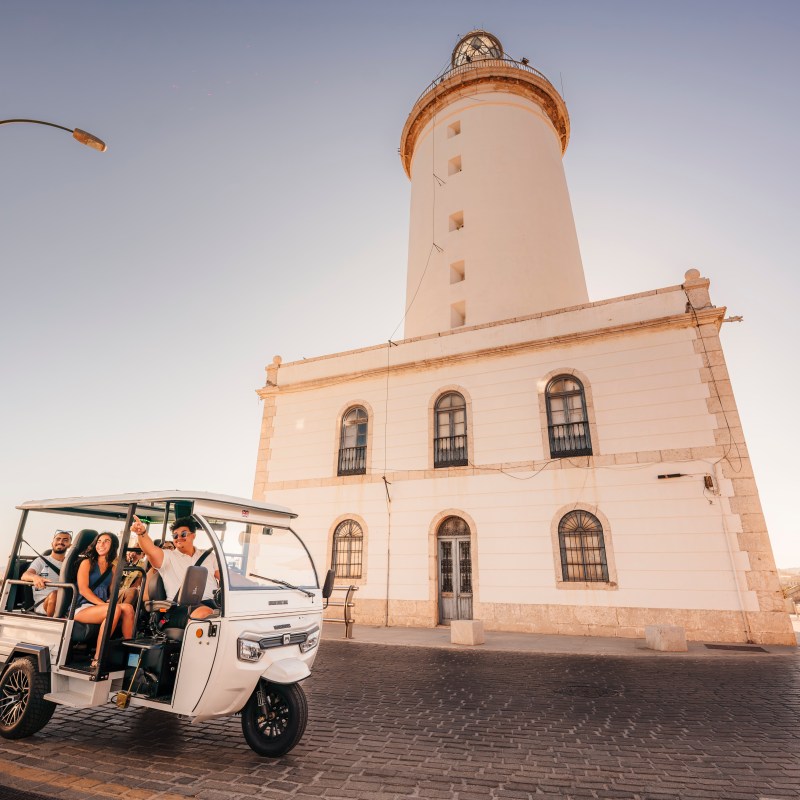 People in a small white vehicle near a lighthouse on a sunny day.