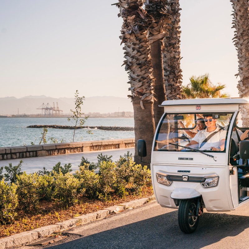 Four people in a white tuk-tuk driving along a seaside road with palm trees and mountains in the background.