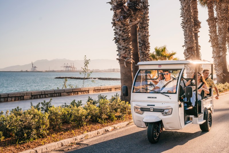 Four people in a white tuk-tuk driving along a seaside road with palm trees and mountains in the background.