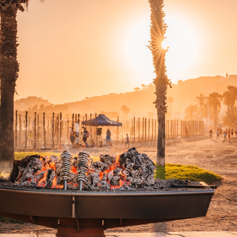 Beachside barbecue at sunset with grilling sardines and palm trees.