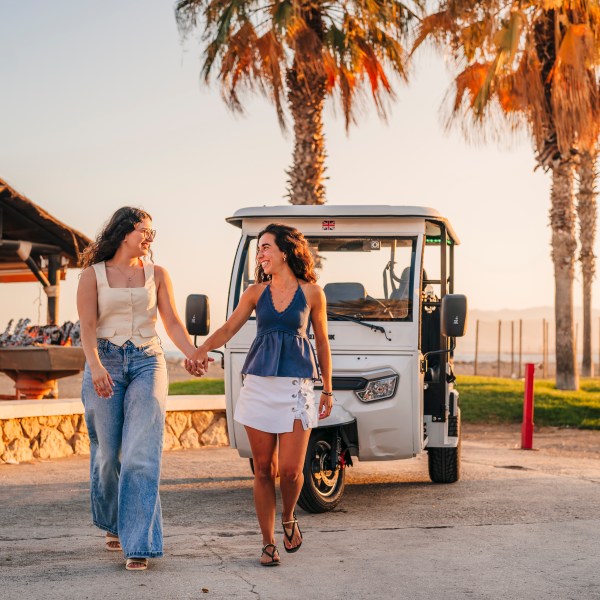Two women holding hands, smiling, walking near a white vehicle with palm trees in the background.