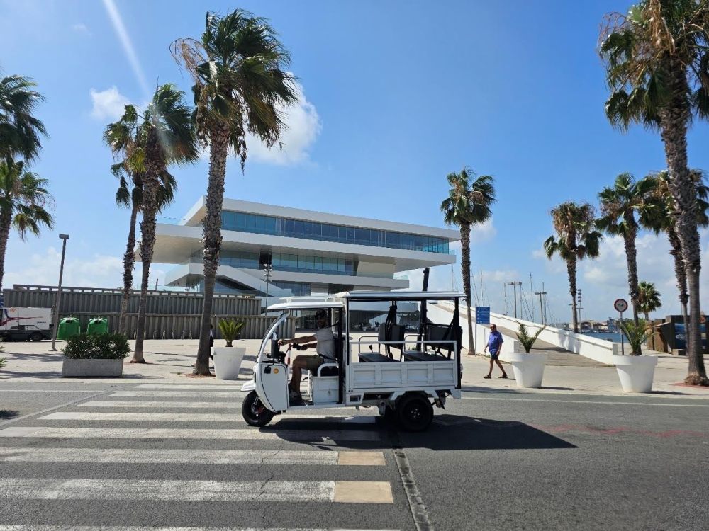 Tuk-tuk eléctrico blanco circulando por la Marina de Valencia frente al edificio Veles e Vents y palmeras bajo un cielo despejado.