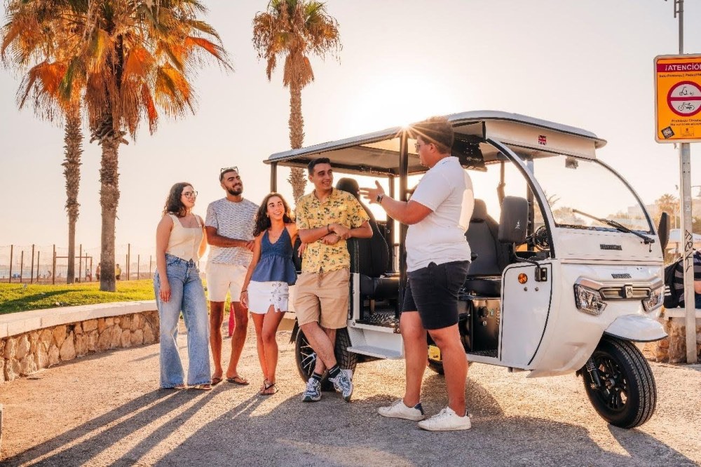 Grupo de amigos escuchando las explicaciones del guía turístico junto a un tuk-tuk eléctrico en el paseo marítimo de Málaga al atardecer