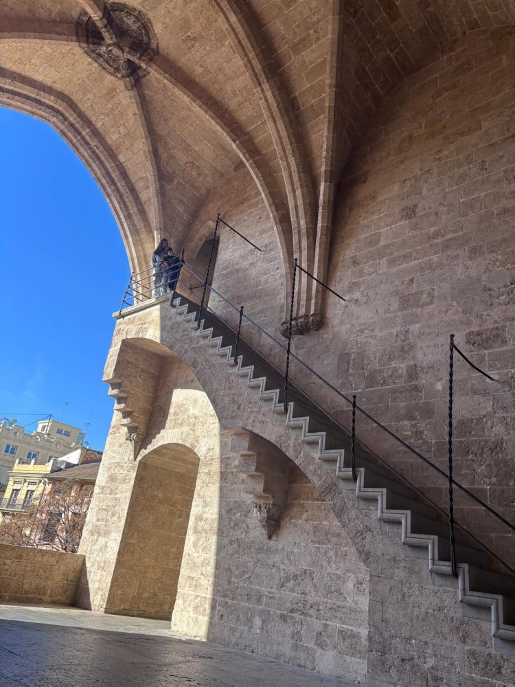 Escalinata de piedra gótica en monumento histórico de Valencia con turistas observando la arquitectura bajo arcos de piedra.