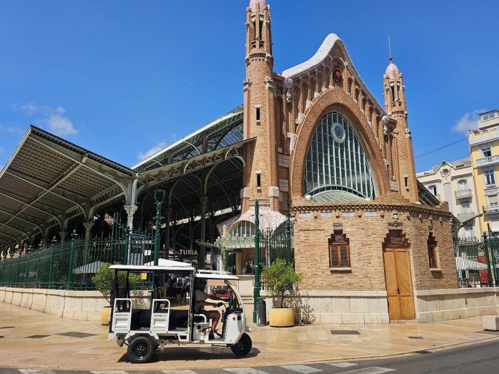 Tuk-tuk eléctrico blanco circulando frente a la fachada modernista del Mercado de Colón en Valencia durante un tour turístico