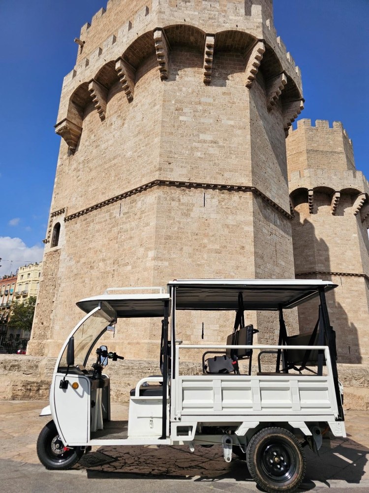 Tuk-tuk eléctrico blanco estacionado frente a las Torres de Serranos en Valencia, arquitectura gótica medieval.