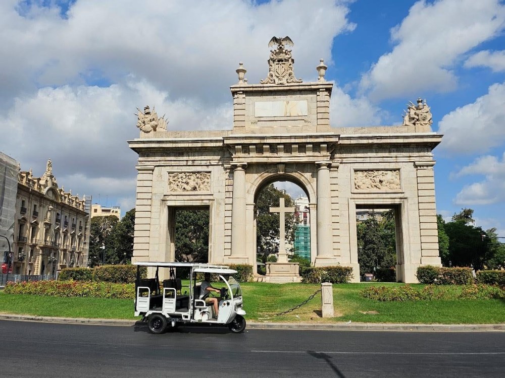 Tuk tuk eléctrico blanco circulando por la Plaza de la Porta de la Mar en Valencia, monumento histórico con arco de triunfo bajo cielo azul.